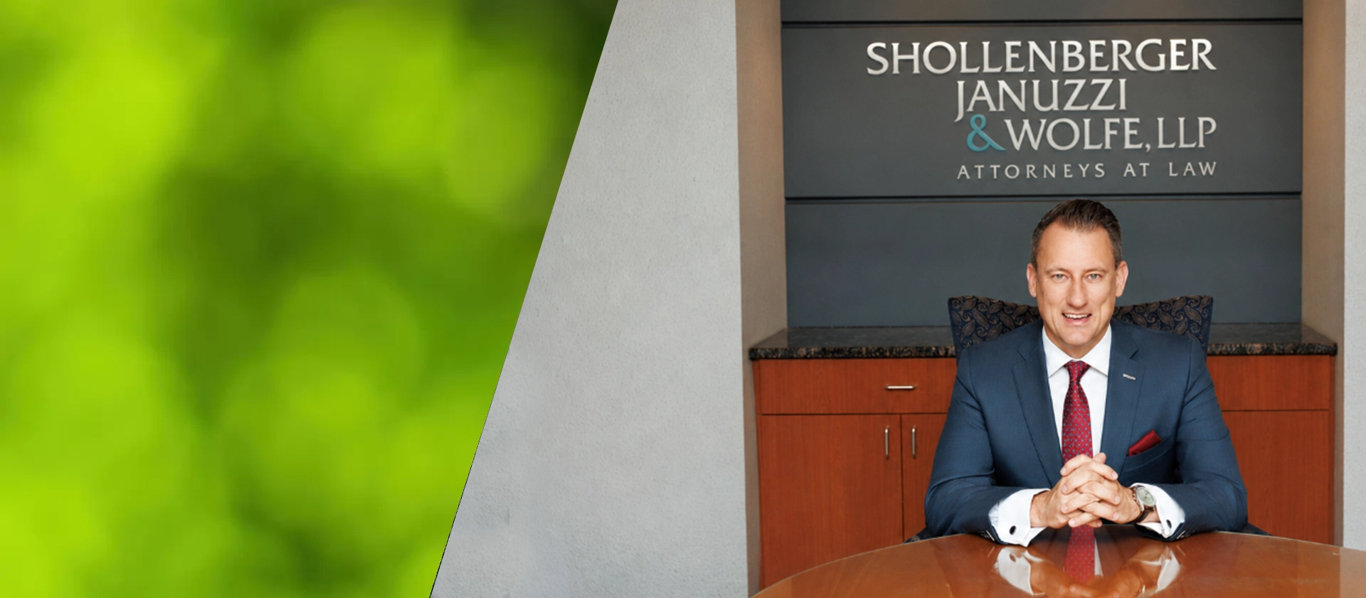 A professional attorney stands next to a dark sign reading 'Shollenberger Januzzi & Wolfe, LLP,' in a modern office environment
